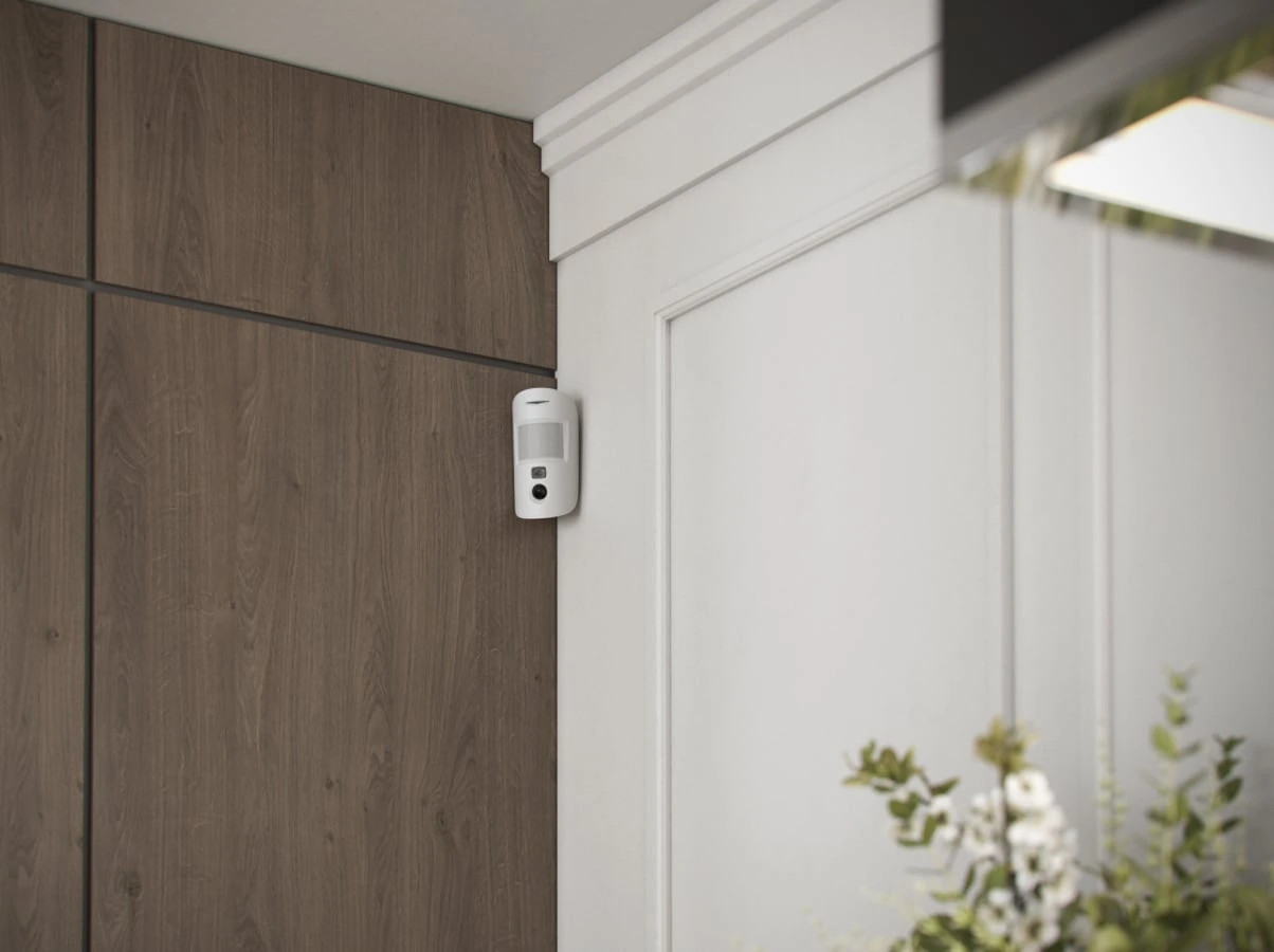 White Ajax MotionCam detector mounted discreetly beside timber wall panels in a finished interior.